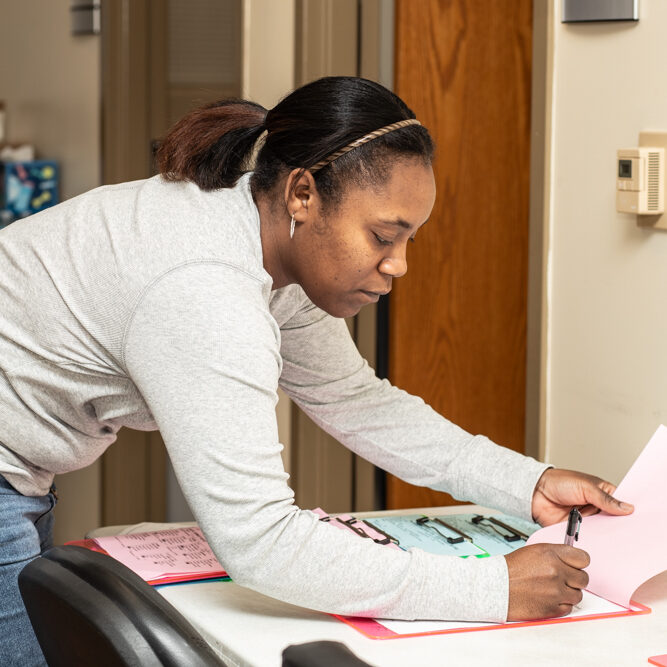 Woman leaning over a table, writing on pink papers with a pen in a well-lit room