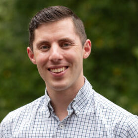 Portrait of a smiling man with short brown hair wearing a white and blue checkered shirt outdoors with greenery in the background.