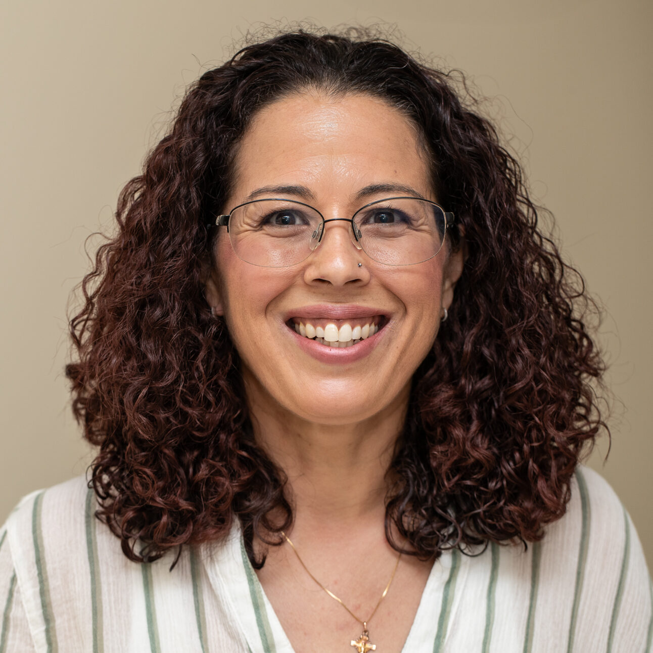 Smiling woman with curly dark hair and glasses, wearing a green-striped white blouse, against a beige background.